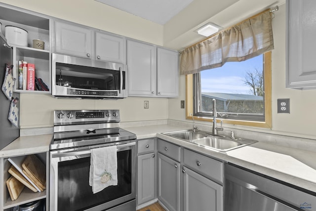 kitchen with a sink, light countertops, appliances with stainless steel finishes, gray cabinets, and open shelves