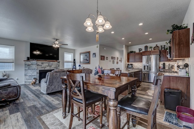 dining area with a textured ceiling, ceiling fan with notable chandelier, wood finished floors, and a stone fireplace