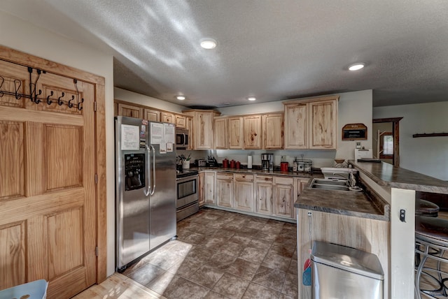 kitchen with dark countertops, appliances with stainless steel finishes, light brown cabinets, a sink, and a peninsula