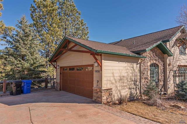 view of side of property featuring an attached garage, stone siding, roof with shingles, and concrete driveway