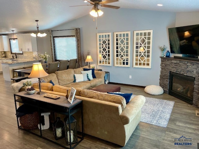 living room with ceiling fan with notable chandelier, vaulted ceiling, wood finished floors, and a stone fireplace