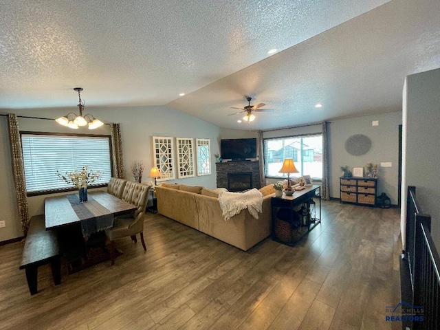 living area with wood finished floors, vaulted ceiling, a textured ceiling, a stone fireplace, and ceiling fan with notable chandelier