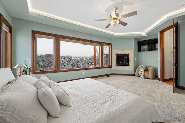 carpeted bedroom with a tray ceiling, a glass covered fireplace, a ceiling fan, and baseboards