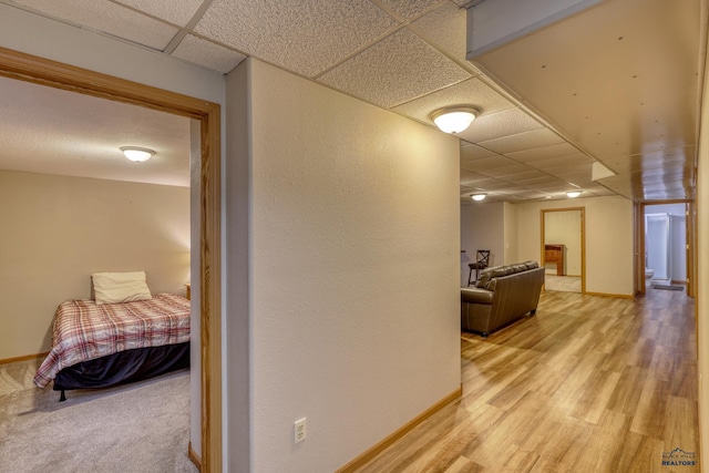 corridor with light wood-type flooring, a paneled ceiling, and baseboards