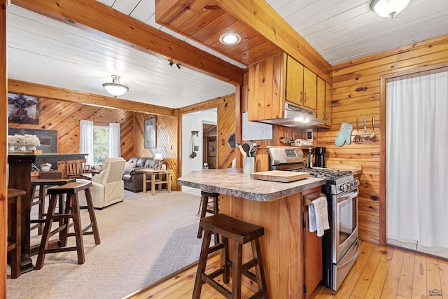 kitchen featuring wooden walls, stainless steel gas range, light wood-type flooring, under cabinet range hood, and beam ceiling