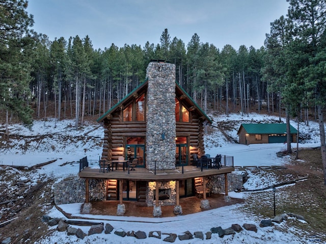 snow covered back of property with a wooden deck, log siding, a forest view, and a chimney