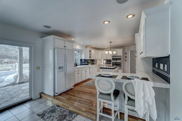 kitchen with white fridge with ice dispenser, a peninsula, white cabinets, decorative backsplash, and tile counters