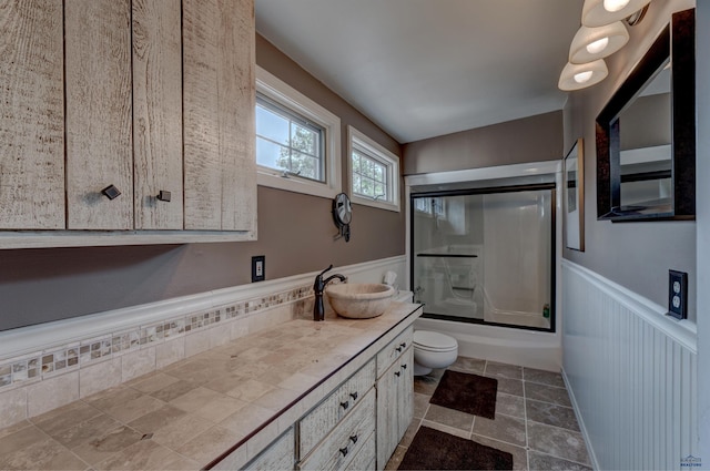 full bath with combined bath / shower with glass door, vanity, toilet, and wainscoting