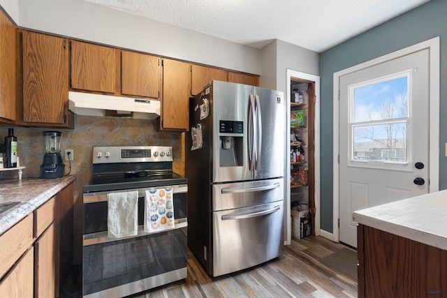 kitchen featuring brown cabinetry, light wood finished floors, stainless steel appliances, under cabinet range hood, and backsplash