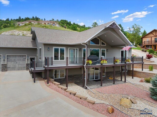 back of property featuring driveway, a shingled roof, stone siding, a garage, and a deck