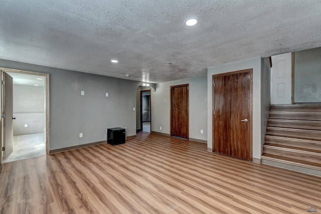 unfurnished living room with a textured ceiling, arched walkways, baseboards, light wood-style floors, and stairway
