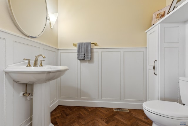 half bathroom with visible vents, a decorative wall, toilet, a wainscoted wall, and a sink