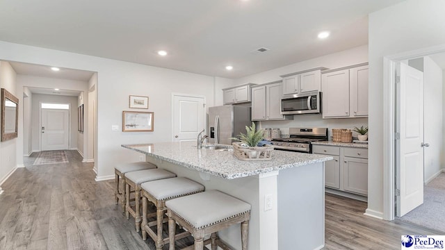 kitchen with gray cabinetry, a center island with sink, a kitchen breakfast bar, stainless steel appliances, and light stone countertops