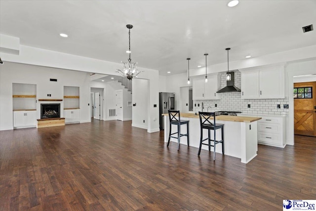 kitchen with butcher block counters, hanging light fixtures, a kitchen island with sink, white cabinets, and wall chimney exhaust hood