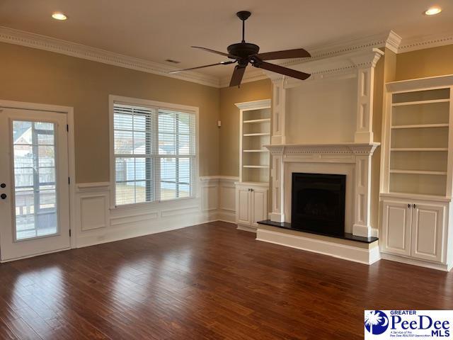 unfurnished living room featuring built in shelves, ornamental molding, dark wood-style floors, wainscoting, and ceiling fan