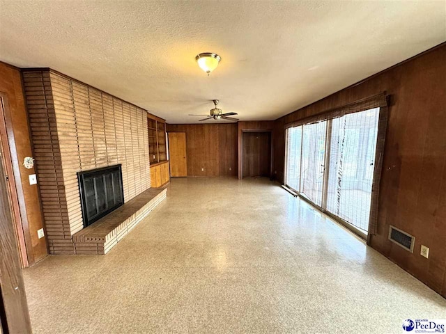 unfurnished living room featuring a textured ceiling, ceiling fan, wood walls, visible vents, and a brick fireplace