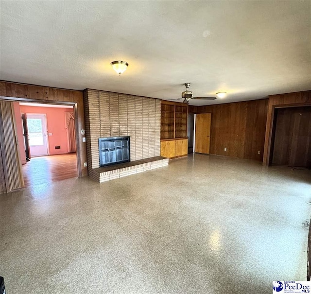 unfurnished living room with a textured ceiling, wooden walls, speckled floor, a ceiling fan, and a brick fireplace