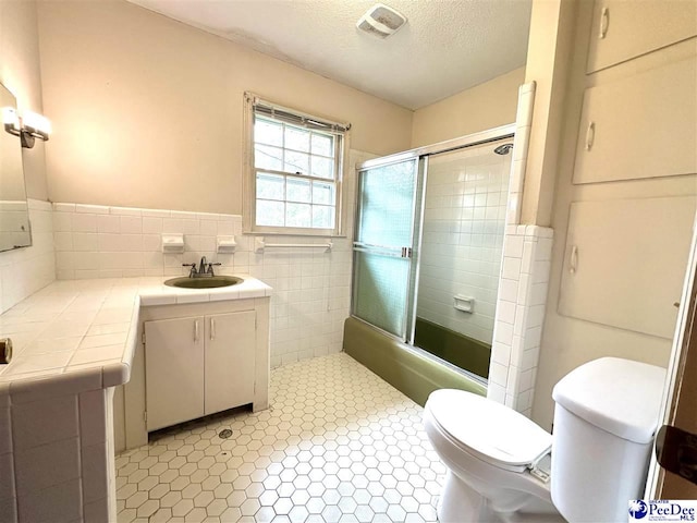 full bathroom with tile walls, shower / bath combination with glass door, vanity, toilet, and a textured ceiling