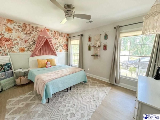 bedroom with wallpapered walls, light wood-type flooring, baseboards, a ceiling fan, and wainscoting