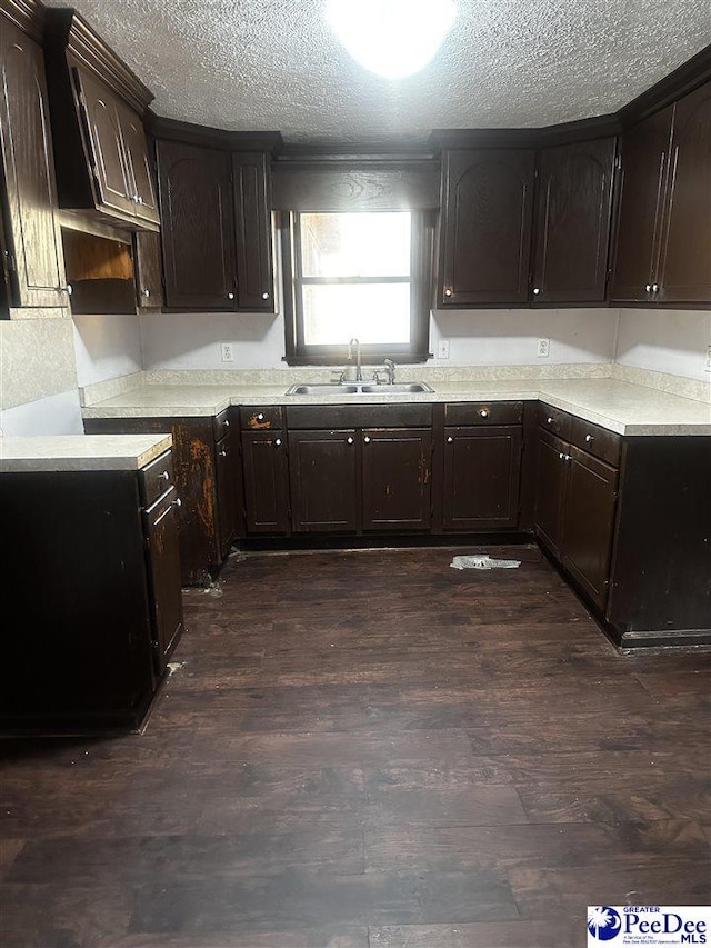 kitchen with a textured ceiling, dark brown cabinetry, a sink, light countertops, and dark wood finished floors