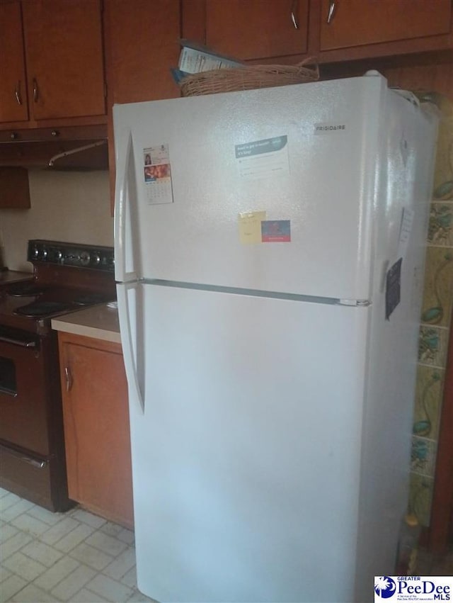 kitchen with under cabinet range hood, electric range, brown cabinetry, and freestanding refrigerator