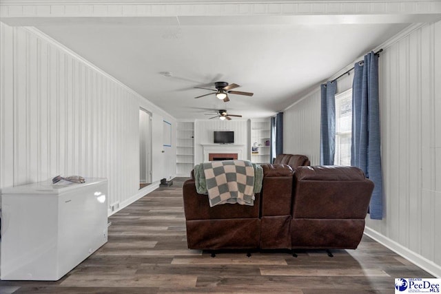 living area featuring built in shelves, a fireplace, a ceiling fan, baseboards, and dark wood finished floors