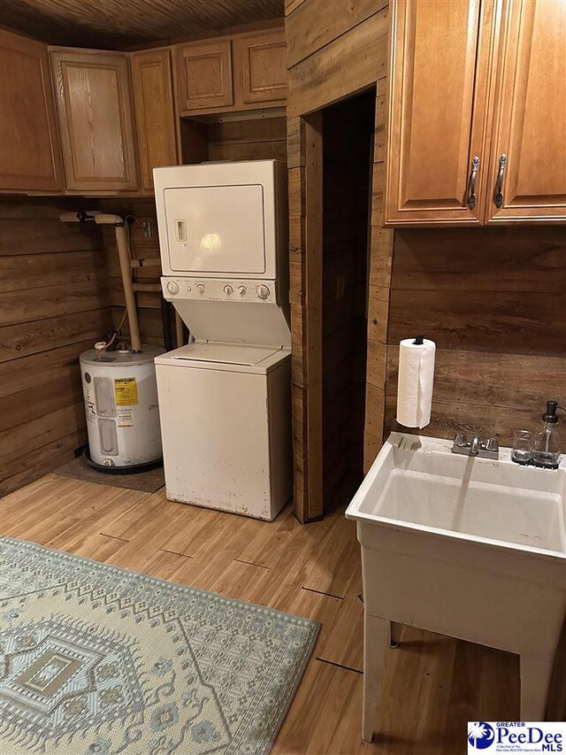 laundry area featuring wooden walls, stacked washer and dryer, cabinets, electric water heater, and light wood-type flooring