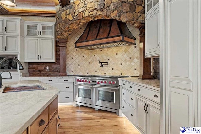 kitchen with sink, premium range hood, light stone counters, light hardwood / wood-style floors, and range with two ovens