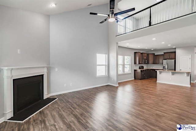 unfurnished living room with ceiling fan, a high ceiling, a fireplace with raised hearth, dark wood finished floors, and baseboards