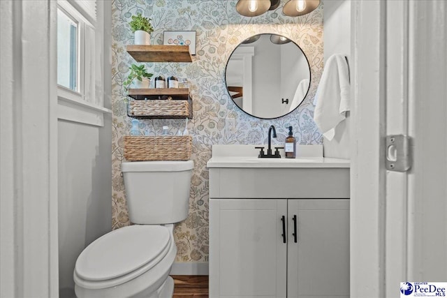 half bathroom with vanity, wood finished floors, baseboards, wallpapered walls, and toilet