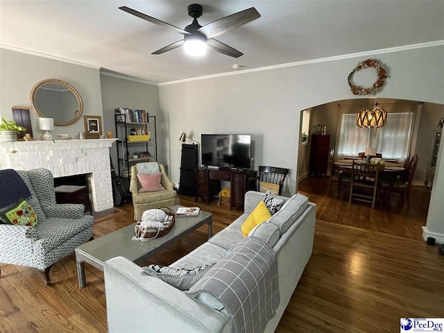 living room featuring crown molding, dark hardwood / wood-style floors, ceiling fan with notable chandelier, and a brick fireplace