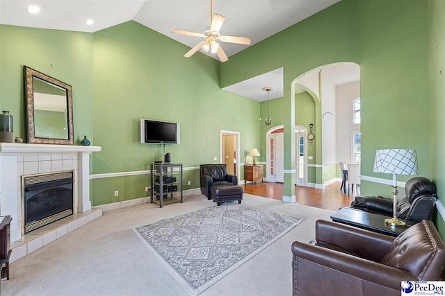 living room with ceiling fan, a tiled fireplace, high vaulted ceiling, and light carpet