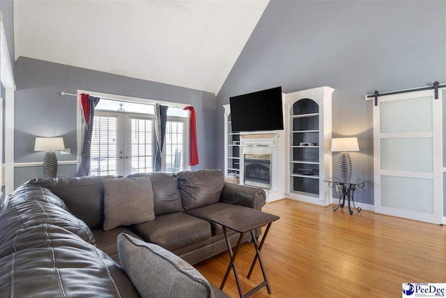 living area featuring a barn door, light wood-style flooring, french doors, a glass covered fireplace, and high vaulted ceiling