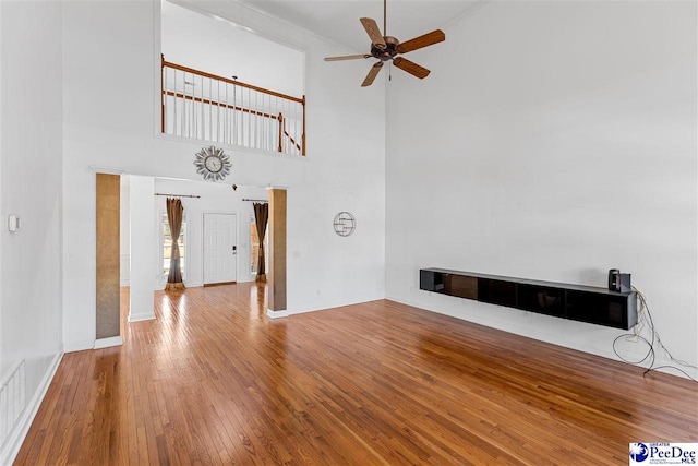 unfurnished living room with ceiling fan, a towering ceiling, visible vents, baseboards, and wood-type flooring