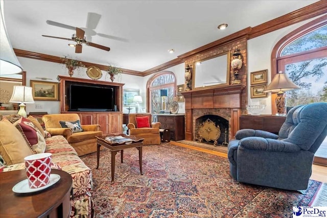 living area with ceiling fan, wood finished floors, a fireplace with raised hearth, and ornamental molding