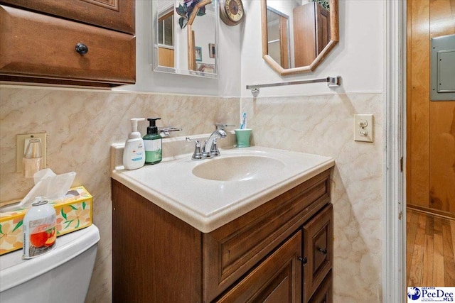 half bathroom featuring tile walls, toilet, wainscoting, vanity, and electric panel