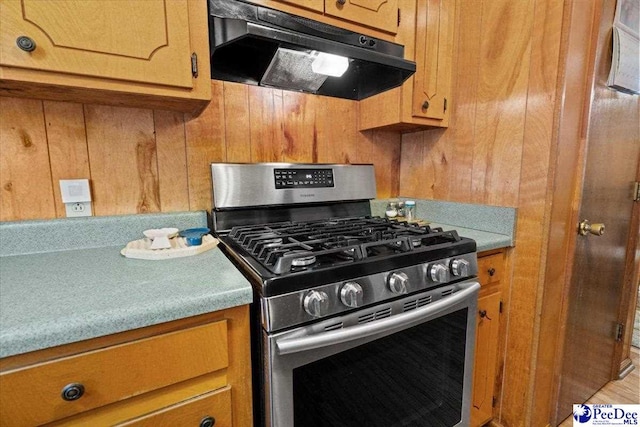 kitchen with wooden walls, under cabinet range hood, light countertops, stainless steel range with gas cooktop, and brown cabinetry