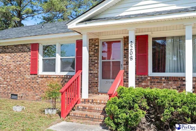 view of exterior entry with a shingled roof, brick siding, and crawl space