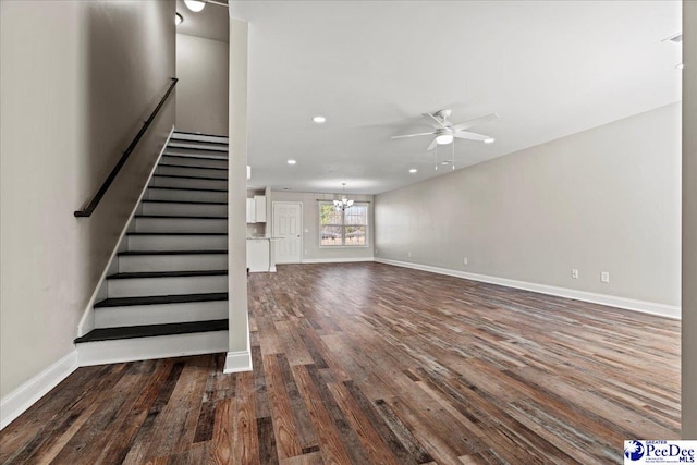 unfurnished living room featuring recessed lighting, ceiling fan with notable chandelier, dark wood-type flooring, baseboards, and stairway