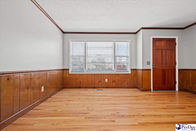 unfurnished room with light wood finished floors, wooden walls, a wainscoted wall, ornamental molding, and a textured ceiling