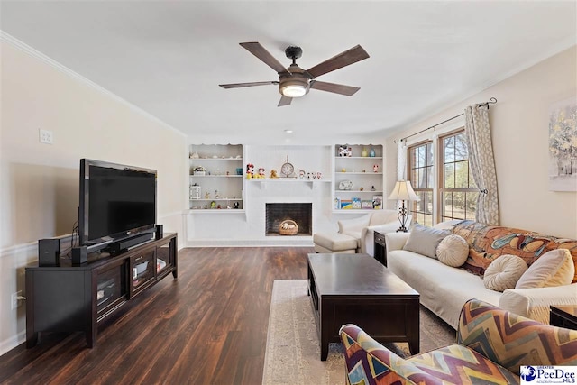 living area featuring built in features, dark wood finished floors, a fireplace with raised hearth, ornamental molding, and ceiling fan