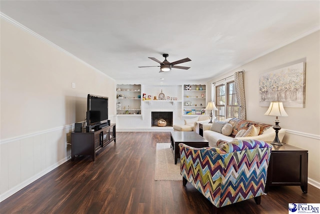 living area featuring built in features, a brick fireplace, wainscoting, dark wood finished floors, and crown molding