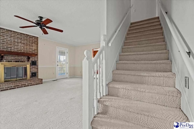 staircase featuring carpet, ornamental molding, a brick fireplace, ceiling fan, and a textured ceiling