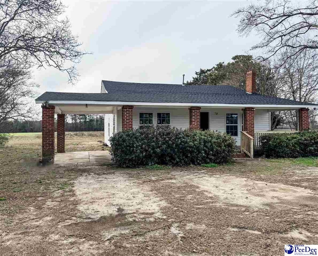 single story home with a shingled roof, brick siding, dirt driveway, a carport, and a chimney