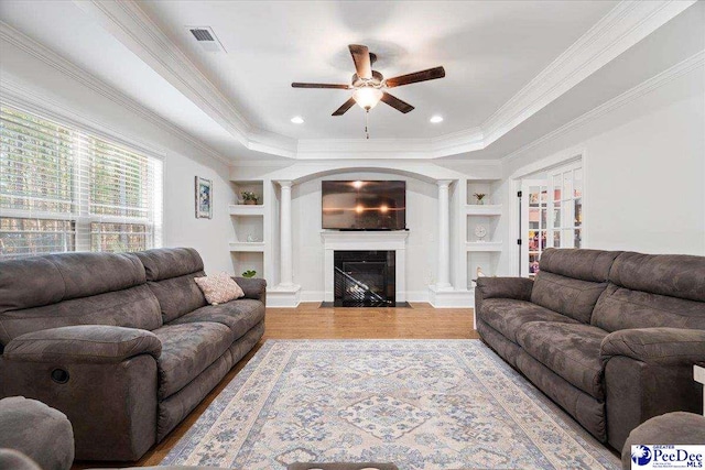 living room featuring built in shelves, crown molding, a raised ceiling, hardwood / wood-style flooring, and decorative columns