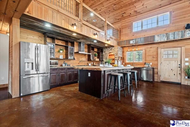 kitchen featuring light countertops, a towering ceiling, appliances with stainless steel finishes, wall chimney range hood, and wooden walls