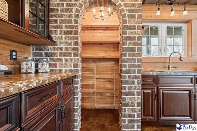 kitchen featuring dark brown cabinetry, glass insert cabinets, stone counters, wood walls, and a sink