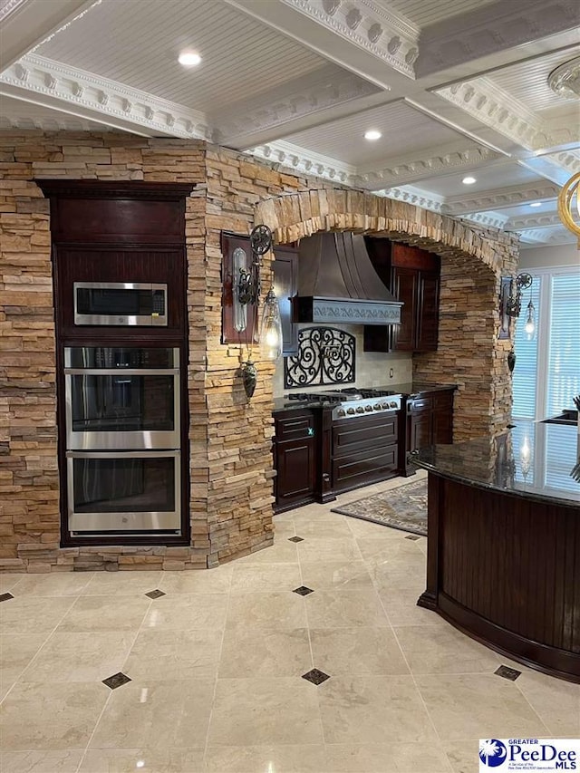 kitchen with dark brown cabinets, stainless steel appliances, coffered ceiling, custom range hood, and beamed ceiling