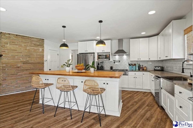 kitchen featuring wall chimney range hood, stainless steel appliances, white cabinets, a kitchen bar, and wood counters