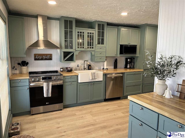 kitchen featuring wall chimney range hood, sink, appliances with stainless steel finishes, green cabinetry, and light wood-type flooring